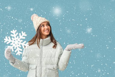 Kışın giyinmiş güzel bir kadın açık mavi arka planda büyük bir kar tanesi tutuyor.