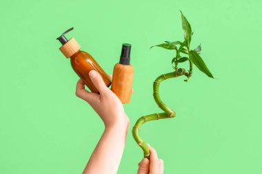 Yeşil arka planda kozmetik ürünlerin şişelerini ve bambu sapını tutan kadın eller