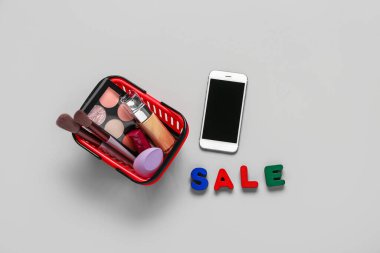 Makyaj malzemeli alışveriş sepeti, modern cep telefonu ve gri arka planda metin satışı.