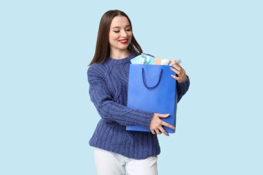 Elinde mavi arka planda Noel hediyesi kutuları olan alışveriş çantası tutan genç bir kadın.