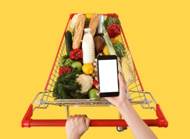 Cep telefonlu bir kadın sarı arka planda yiyecek dolu alışveriş arabalarını itiyor.