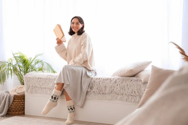 Sıcak kazaklı genç bir kadın evde kitap okuyor.