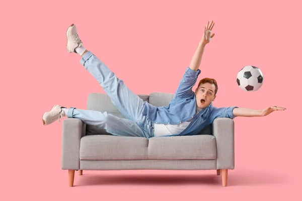 Young man with soccer ball lying on grey sofa against pink background