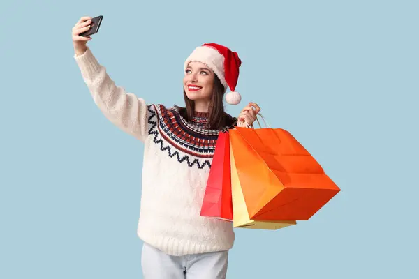 サンタハットの若い女性は 青い背景にショッピングバッグでセルフィーを取る — ストック写真