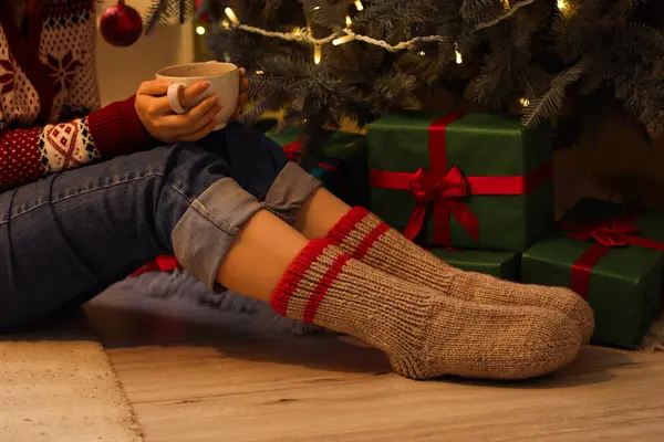 Woman in warm socks with cup of tea at home near Christmas tree