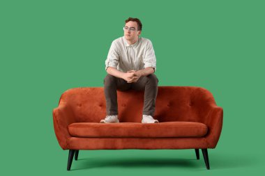 Genç adam yeşil arka planda kırmızı koltukta oturuyor.