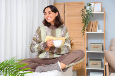 Evde bir fincan çay ve kitapla sıcak kazak giyen genç bir kadın.