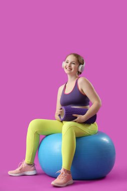 Köpüklü silindirli ve kulaklıklı güzel genç bir kadın pembe arka planda fitness topunun üzerinde oturuyor.