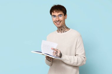 Mavi arka plan üzerine kitabı olan dövmeli genç bir adam.