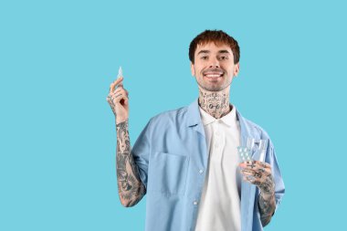 Mavi arka planda burun damlası kullanan ve bir bardak su içen alerjisi olan genç bir adam.