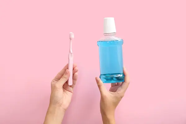 Female hands with toothbrush and mouth rinse on pink background.
