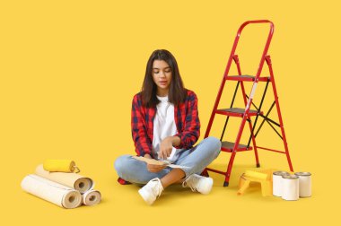 Sarı arka planda oturan boya rengi paletleri ve aletleri olan genç bir kadın.