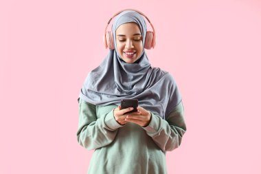 Kulaklıklı genç Müslüman kadın pembe arka planda cep telefonu kullanıyor.