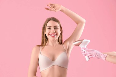 Güzel genç bir kadın ve elleri pembe arka planda modern fotoepilatörle koltukaltlarına epilasyon yapıyor.