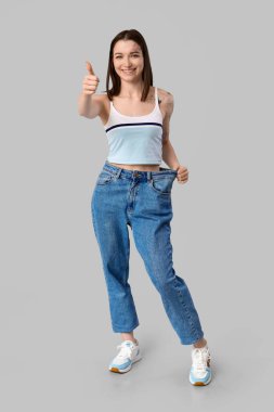 Gevşek kotlu güzel genç bayan gri arka planda baş parmak hareketi gösteriyor. Kilo verme konsepti