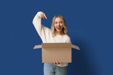 Genç bir kadın mavi arka plandaki açık karton kutuyu işaret ediyor.
