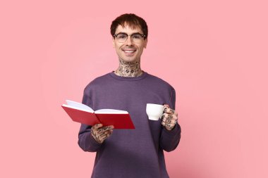 Pembe arka planda kitap ve bir fincan kahve olan dövmeli genç bir adam.