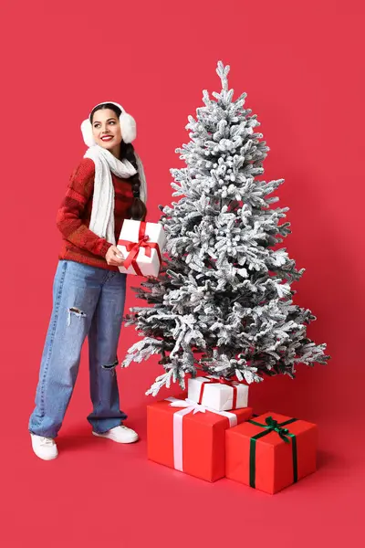 赤い背景にクリスマスツリーの近くにギフトボックスを持つ美しい若い女性 — ストック写真
