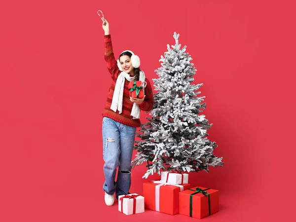 红底圣诞树旁 一位漂亮的年轻女子拿着糖果手杖和礼品盒 — 图库照片