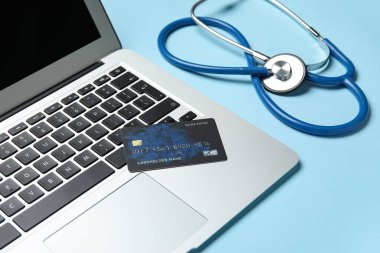 Mavi arka planda dizüstü bilgisayar, kredi kartı ve steteskop.