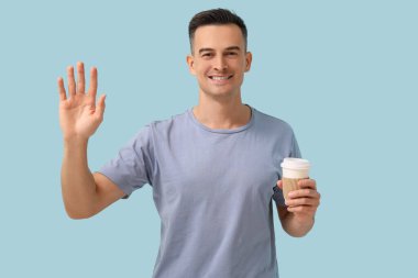 Elinde kahve fincanı olan yakışıklı bir adam mavi arka planda el sallıyor.