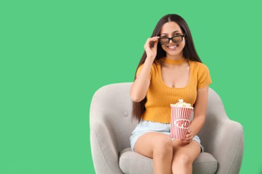 3 boyutlu gözlüklü ve patlamış mısırlı genç bir kadın yeşil arka planda koltukta film izliyor.