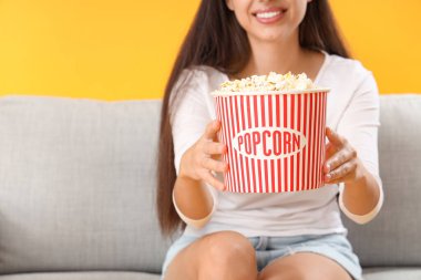 Patlamış mısırlı genç bir kadın kanepede sarı arka planda film izliyor.