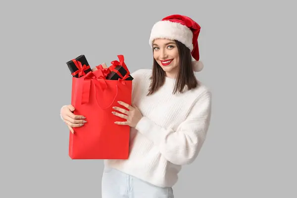 サンタハットの若い女性は 灰色の背景にギフトボックスを持つショッピングバッグを保持しています — ストック写真
