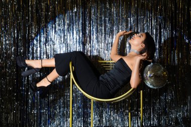 Elinde disko topu olan genç bir kadın gümüş süslemelere karşı koltukta oturuyor.