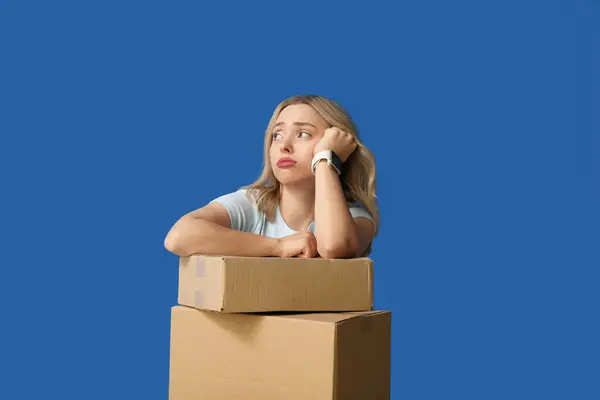 Sad young woman with cardboard boxes on blue background