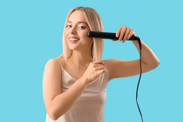 Young blonde woman straightening hair on blue background