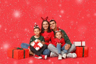 Mutlu aile Noel süveterleri içinde ve kırmızı arka planda hediyeleri var.