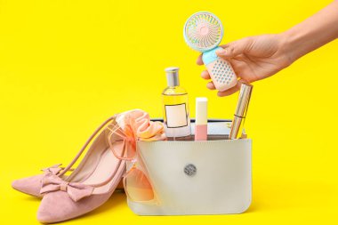 Taşınabilir elektrikli vantilatörü ve sarı arka planda makyaj malzemeleri olan bir kadın eli.