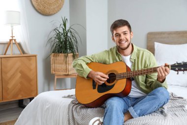 Genç sakallı adam yatak odasında gitar çalıyor.