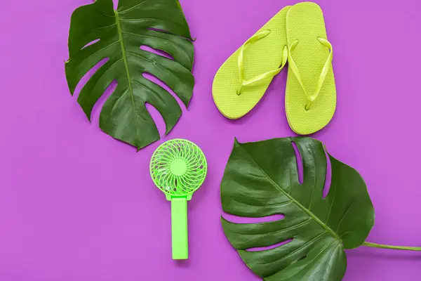 Portable green electric fan, flip-flops and monstera leaves on purple background