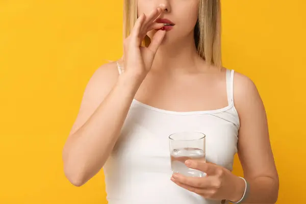 Young woman with vitamin A pill and glass of water on yellow background, closeup