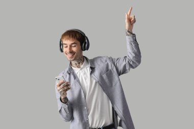 Gri arka planda kulaklık ve cep telefonuyla müzik dinleyen mutlu genç adam.