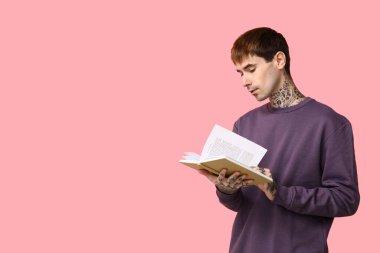 Genç dövmeli adam pembe arka planda kitap okuyor.