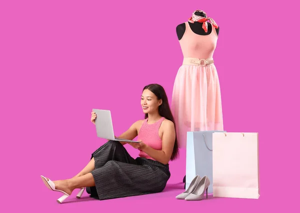 Female Asian stylist with laptop, shopping bags and mannequin on purple background