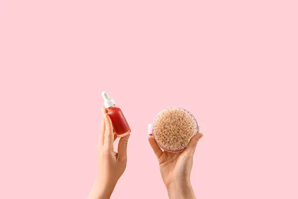Female hands holding scrubbing brush and bottle of cosmetic product on pink background