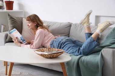 Evdeki kanepede dergi okuyan genç bir kadın.