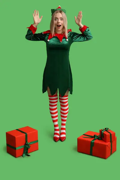 Surprised young woman in elf costume with Christmas gift boxes on green background