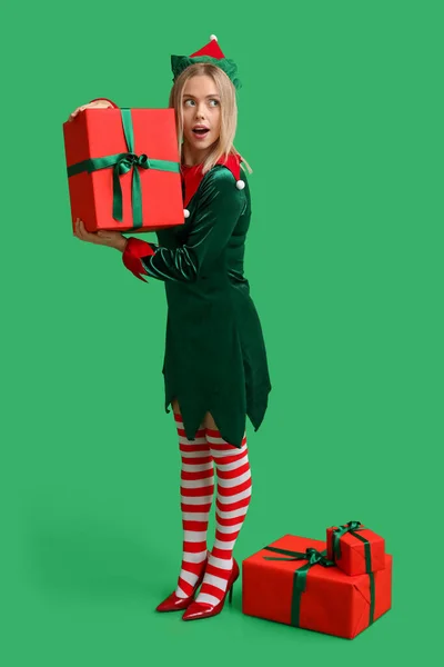 Emotional young woman in elf costume with Christmas gift boxes on green background