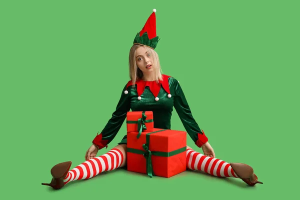Young woman in elf costume with Christmas gift boxes on green background