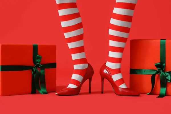 Legs of young woman in elf's stockings with Christmas gift boxes on red background