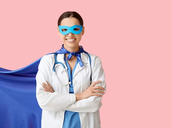 Young female doctor in superhero costume on pink background