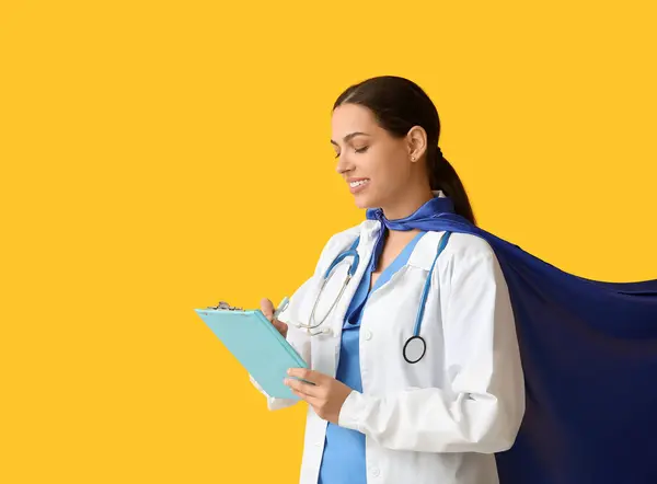 Beautiful female doctor in superhero costume with clipboard on yellow background