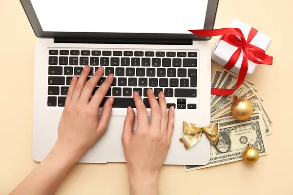 Female hands with laptop, money and Christmas gift box on yellow background