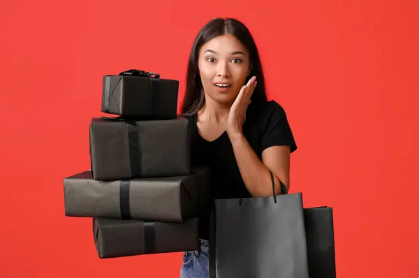 Shocked young Asian woman with gift boxes and shopping bags on red background. Black Friday sale