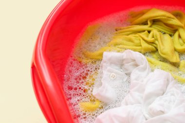 Kirli elbiseli plastik leğen, sarı arka planda su ve köpük, yakın plan.